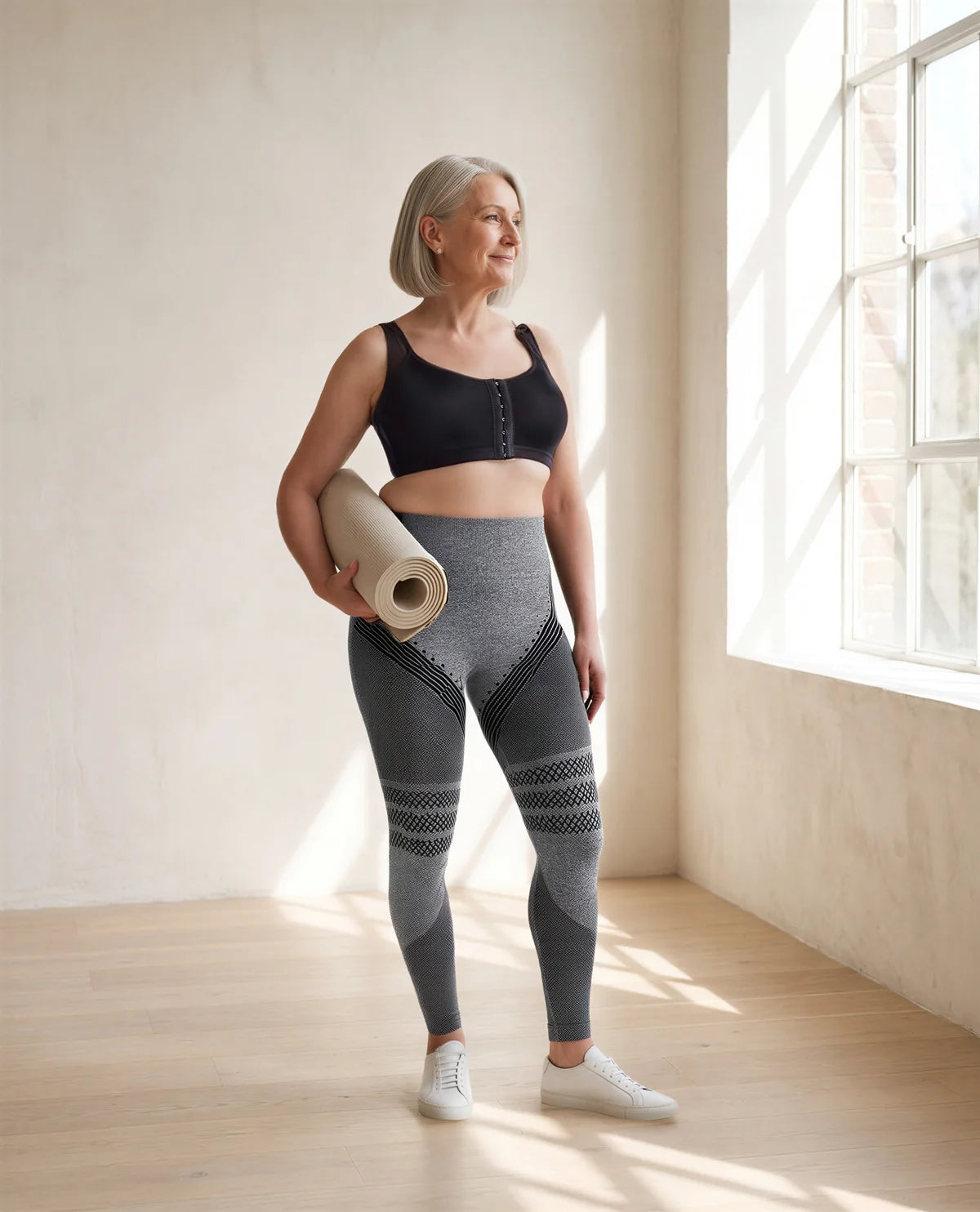 A woman in her fifties carrying a yoga mat, in the black bra and grey leggings