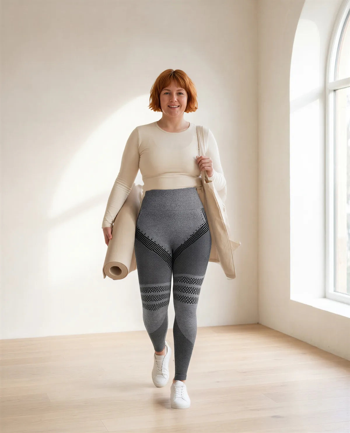 A woman with a rolled yoga mat walking across a sunlit studio in heather grey Fairhold leggings