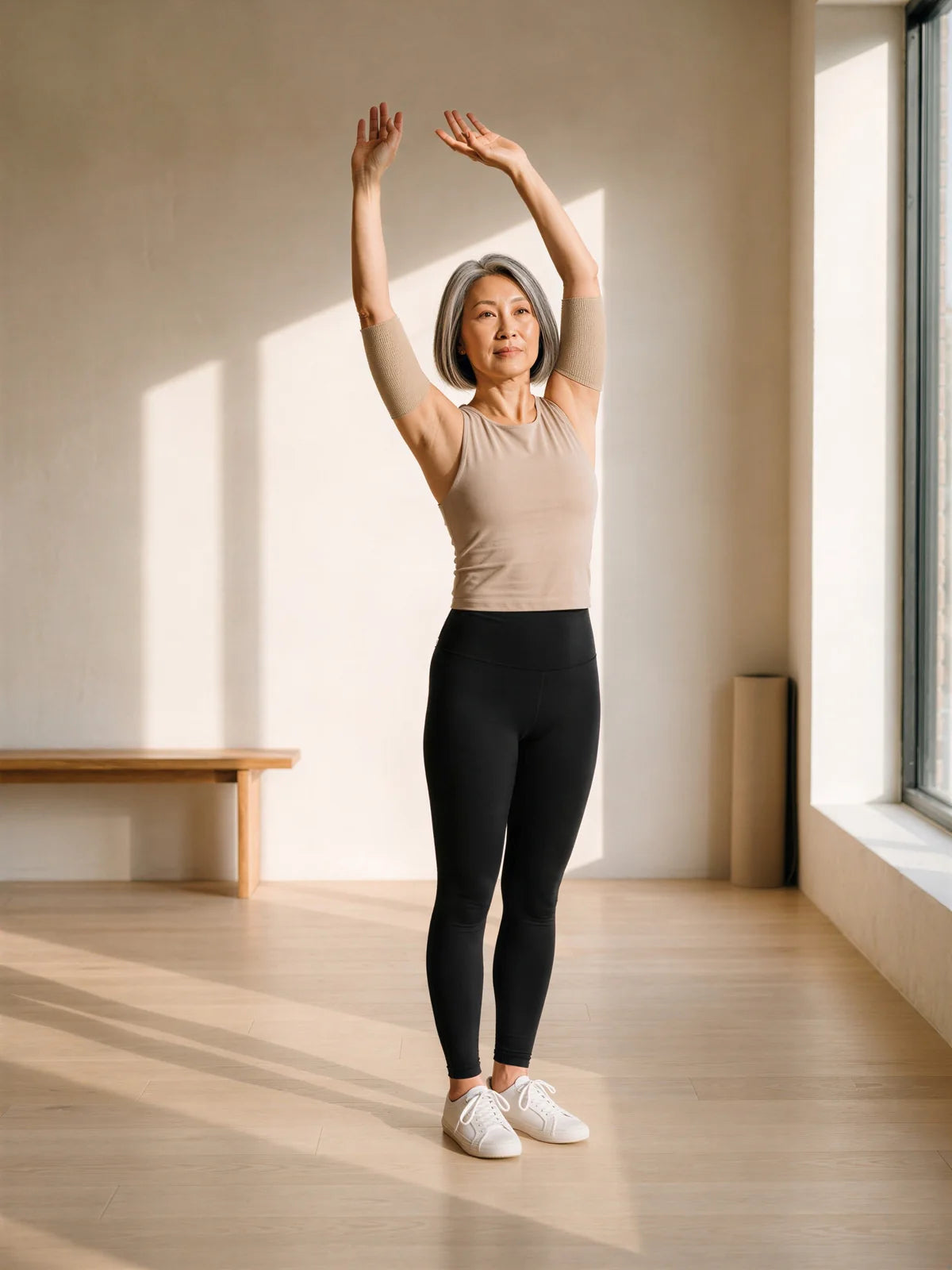 A woman in her fifties in a slow overhead stretch, wearing beige arm sleeves