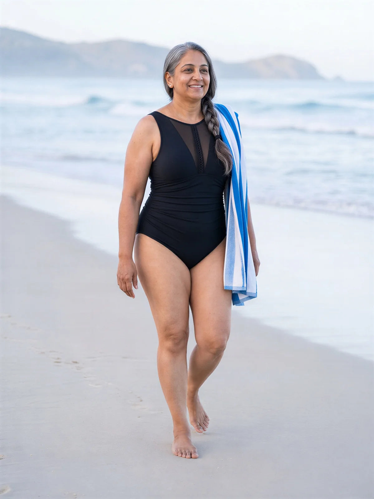 A woman in her mid fifties walking along the beach with a towel, in the black one-piece